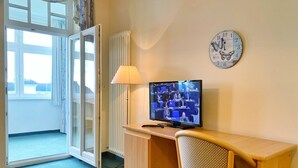 Una habitación con un escritorio de madera, un televisor mostrando una reunión, un reloj y una ventana con cortinas.