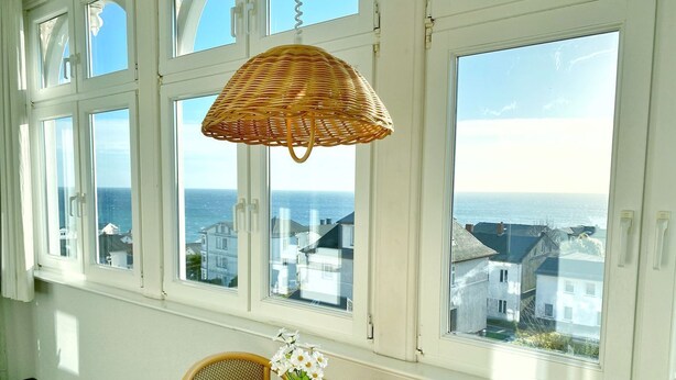 A room with large windows overlooking a coastal town, a wicker lamp, a table with a vase of flowers, and wicker chairs. - Sea view apartment no. 22 in the Villa Anna in Sassnitz on Rügen - Sassnitz - (Sassnitz)