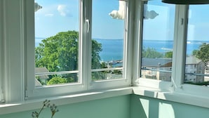 Ein Zimmer mit Blick auf das Meer und Bäume, ein weißer Stuhl und eine Vase mit violetten Blumen.