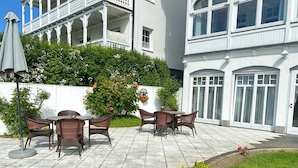 Eine Terrasse im Freien mit Korbmöbeln, ein weißes Gebäude mit Balkon und ein Gartenbereich mit Blumen.