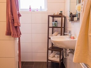 Un baño con un lavabo blanco, un estante de madera con artículos de aseo y una ventana sobre el lavabo.