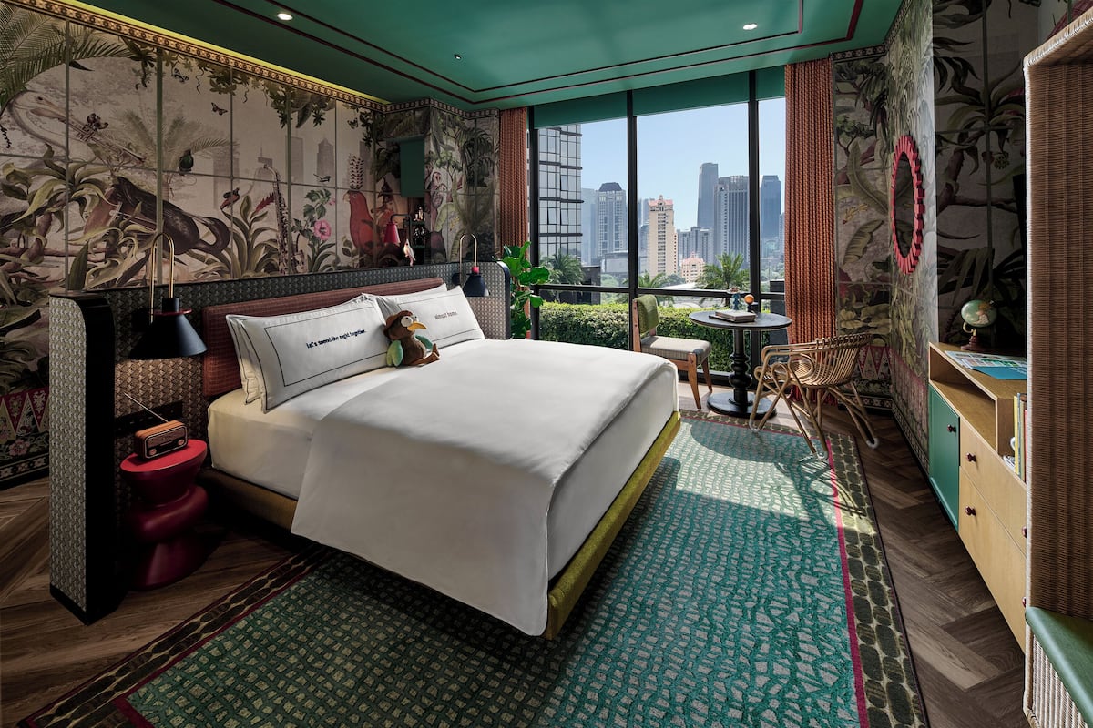 A hotel room with a large bed, a small table, a chair, and a view of the cityscape through large windows.