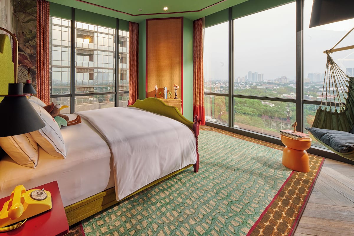 A hotel room with a large bed, a red side table, a yellow rotary phone, and a view of a cityscape through large windows.