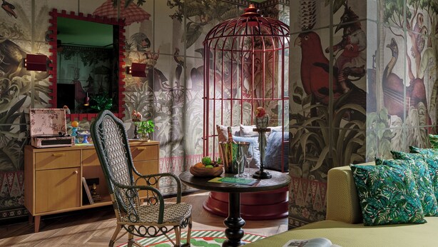 Una habitación con una silla de estilo antiguo, una mesa redonda con un globo terráqueo, una estantería y un ventanal con vistas a una jaula de pájaros.