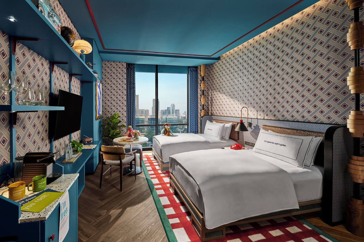 A hotel room with two beds, a desk with a computer, a chair, a small table with a plant, and a view of the city skyline.