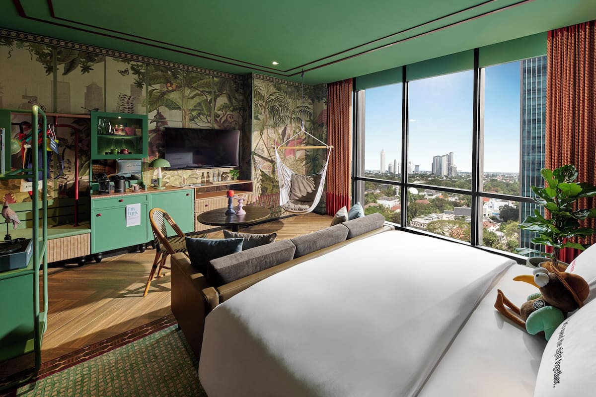 A hotel room with a large bed, a desk with a TV, a seating area with a view of the city, and a green-themed decor.
