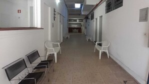 A long, narrow hallway with white walls and tiled floors, lined with white plastic chairs and a row of black metal benches on one side. - Pousada Baraúna (Santos)