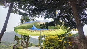 A table and two chairs under a green umbrella, surrounded by trees and a brick wall. - Remarkable 1-Bed House in Bhimtal (Bhimtal)