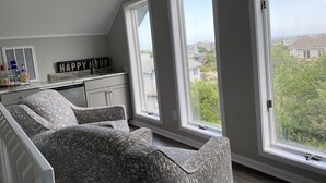 A room with a sloped ceiling, a large window, a sofa, a kitchen area with cabinets, and a sign that reads "HAPPY HOUR". - Oceanside DUCK Beach Home, 5 minute walk to Beach & Shops (Duck)