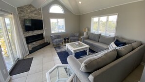 A spacious living room with a stone fireplace, a ceiling fan, and a large TV. - Oceanside DUCK Beach Home, 5 minute walk to Beach & Shops (Duck)