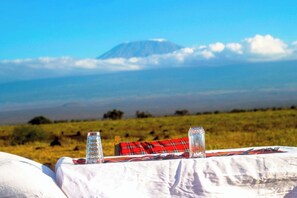 Una mesa con mantel a cuadros rojo y blanco, dos vasos transparentes y una botella, con un campo cubierto de hierba y una montaña de fondo.