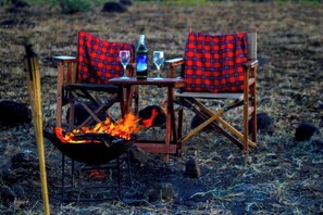 A campfire with two plaid chairs and a table with a bottle and glasses.