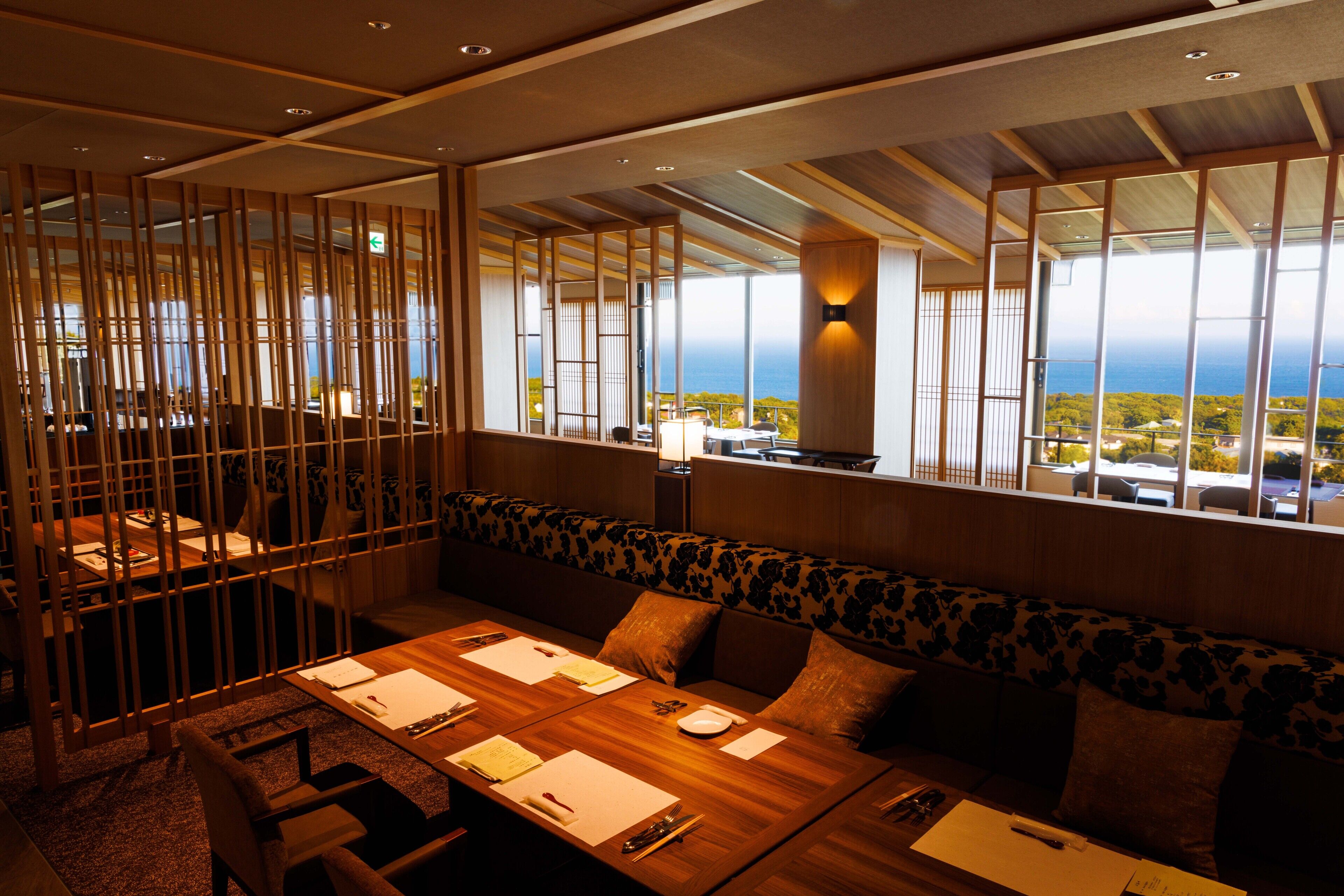 A dining area with a long wooden table set for a meal, a sofa with floral upholstery, and large windows offering a view of the ocean.