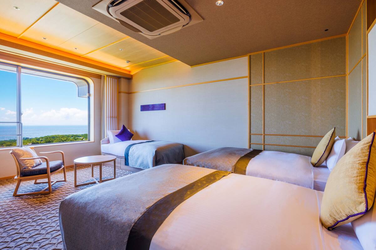 Une chambre d’hôtel avec deux lits, un canapé, un fauteuil et une grande fenêtre donnant sur la mer.