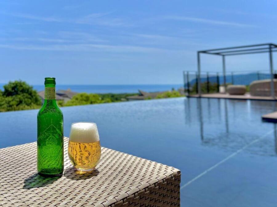 Une bouteille verte et un verre de bière sont posés sur une table près d’une piscine, avec vue sur l’océan.