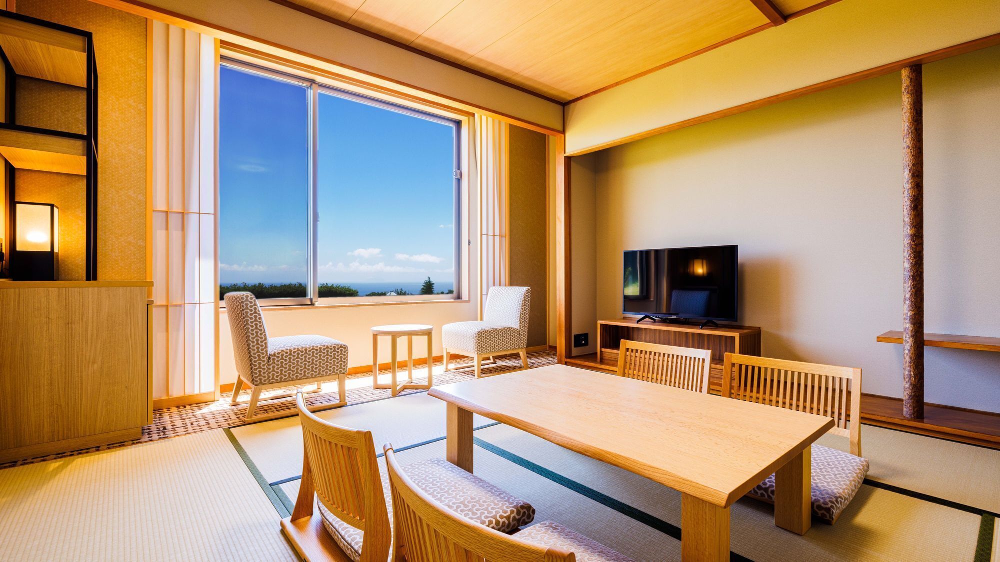 A traditional Japanese-style room with tatami flooring, a low table, chairs, a television, and a large window offering a view of the outdoors.