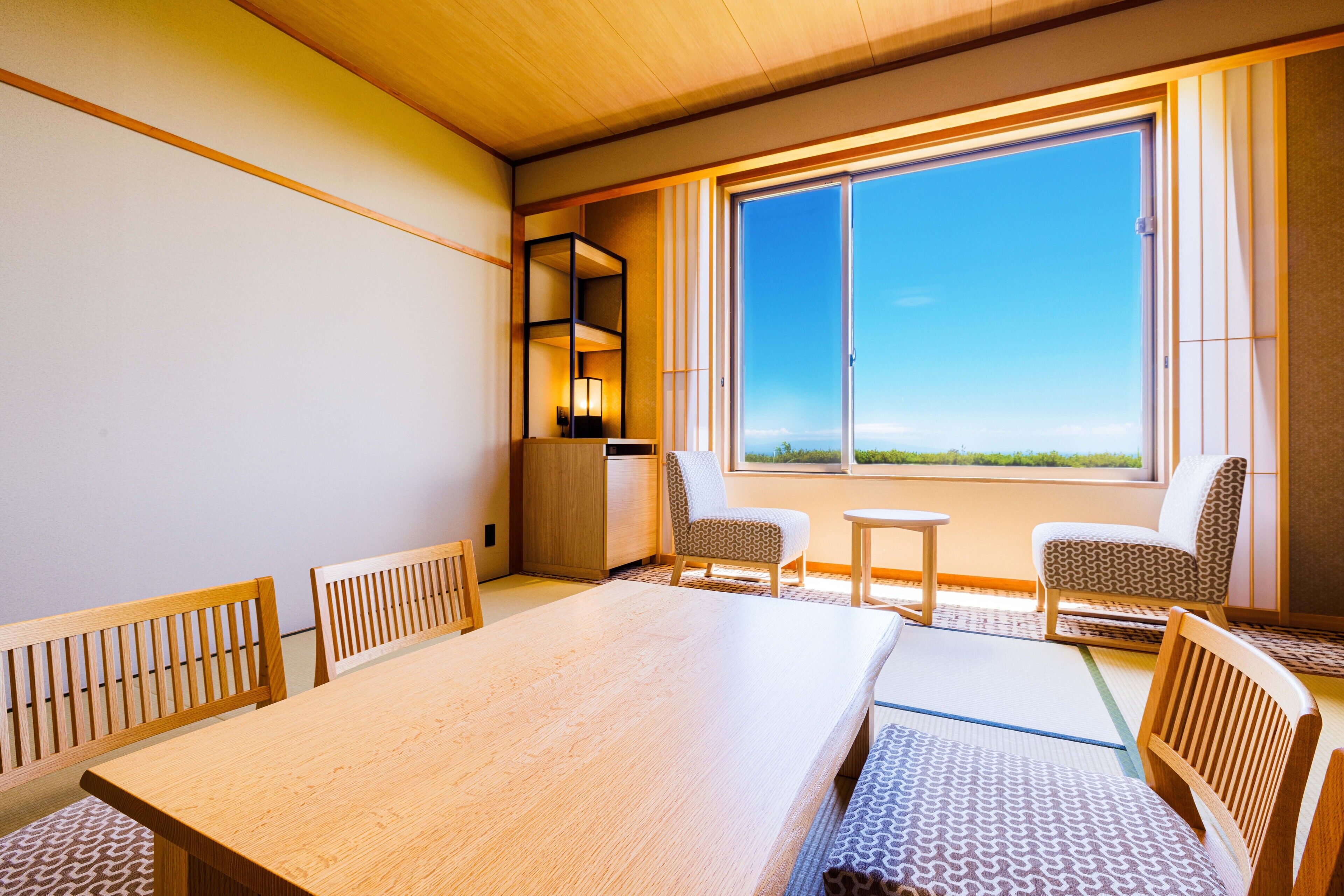 Une chambre d’hôtel avec une grande table en bois, deux chaises à motifs, une petite table et une grande fenêtre donnant sur un beau paysage.