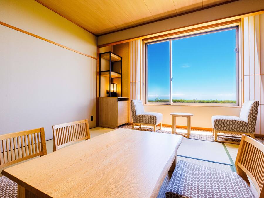 Une chambre d’hôtel avec une grande table en bois, deux chaises à motifs, une petite table et une grande fenêtre donnant sur un beau paysage.