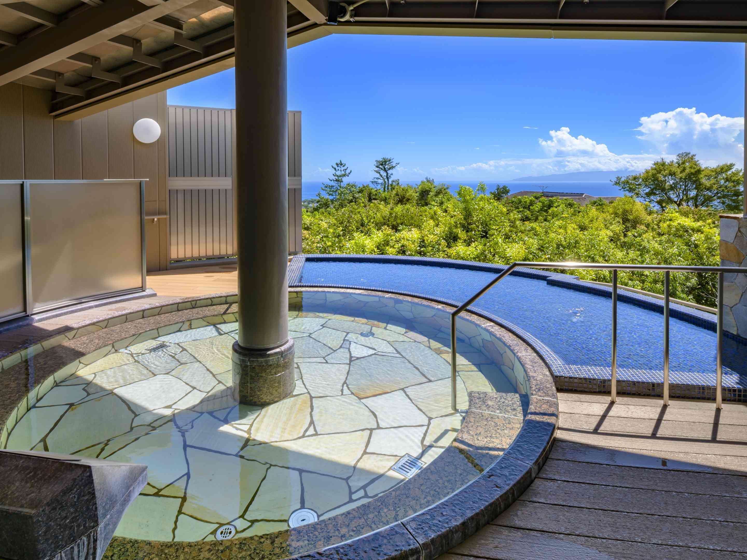 Ein Whirlpool mit Steinmosaik-Design, umgeben von einer Holzterrasse und einem Metallgeländer, mit Blick auf eine üppige grüne Landschaft und den Ozean.