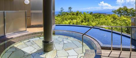 Ein Whirlpool mit Steinmosaik-Design, umgeben von einer Holzterrasse und einem Metallgeländer, mit Blick auf eine üppige grüne Landschaft und den Ozean.