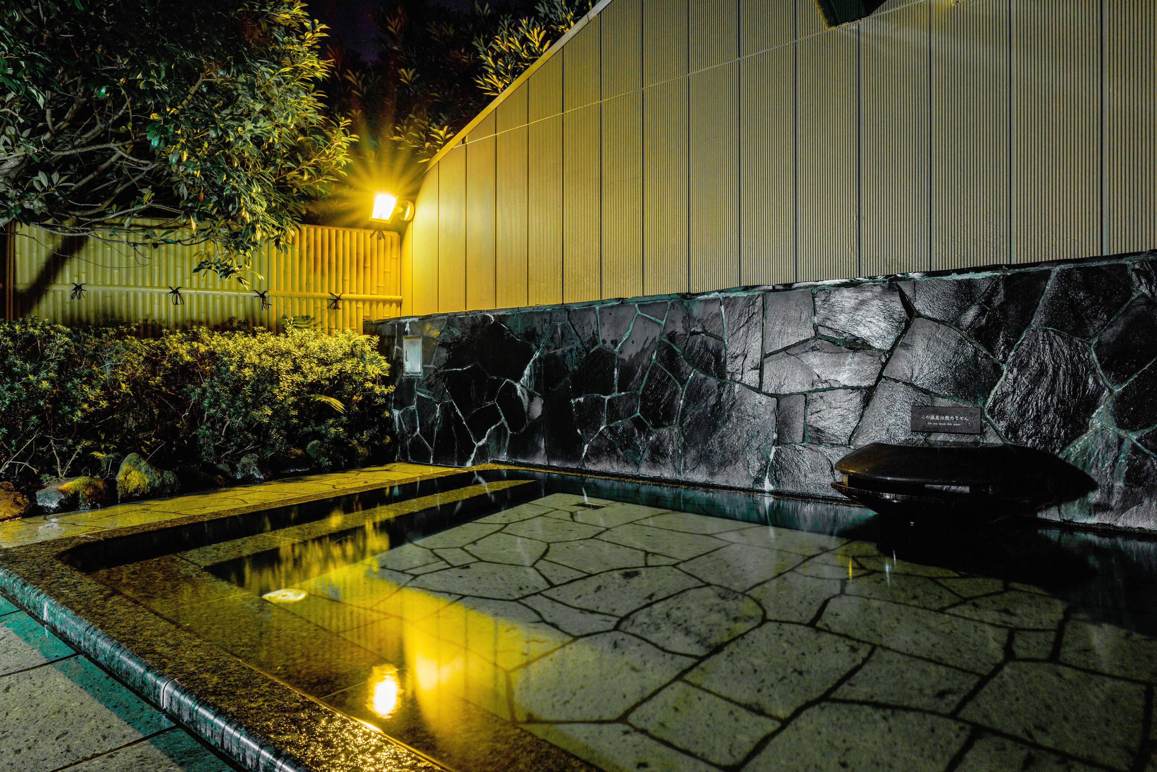 Un área de piscina exterior de noche con baldosas de piedra, una valla metálica y una pared con líneas verticales.