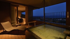 Un balcón moderno con un jacuzzi, una silla de descanso y vistas a la ciudad al anochecer.