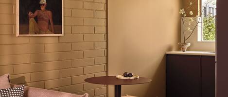 A cozy living room with a brick wall, a painting, a round table with chairs, a sofa, and a small kitchen area with a window.