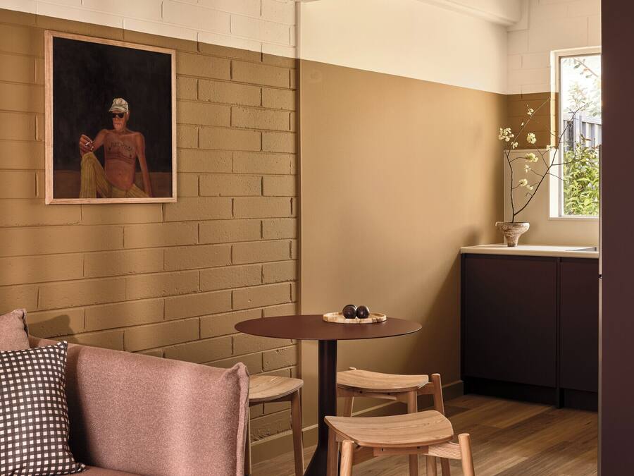 Un salon chaleureux avec un mur en briques, un tableau, une table ronde entourée de chaises, un canapé et un petit coin cuisine avec une fenêtre.