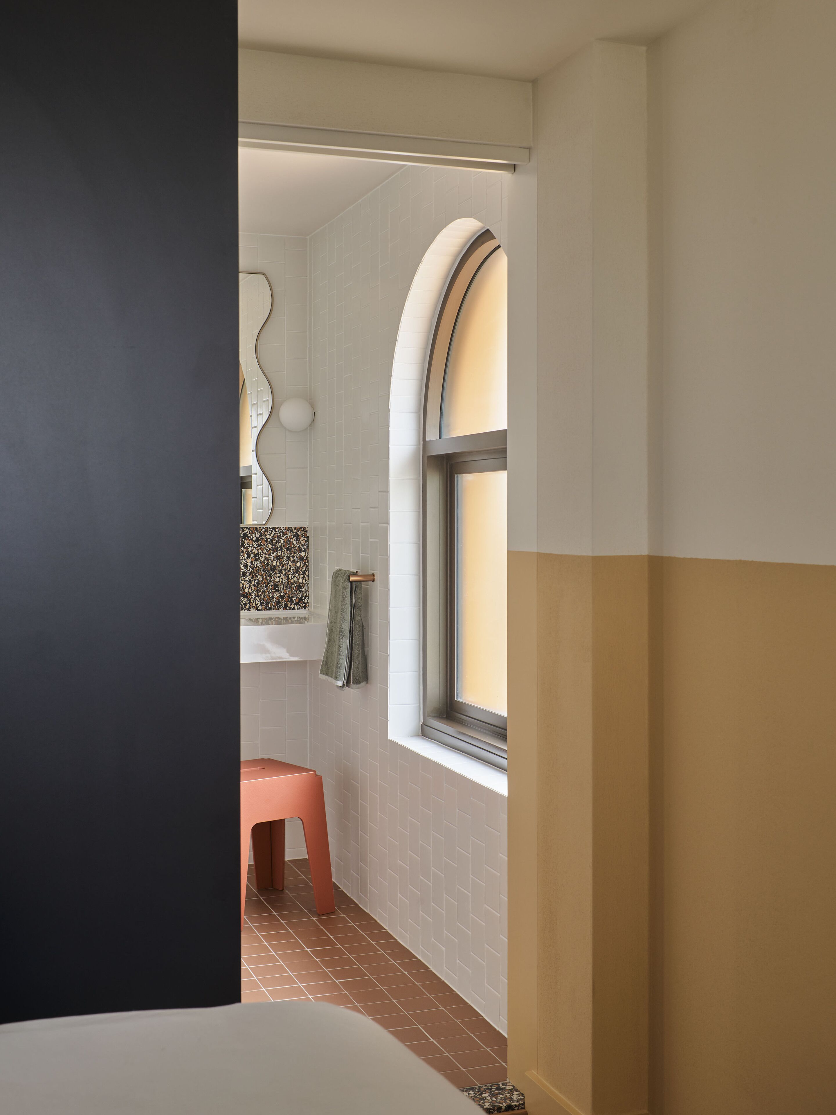 Een badkamer met een witte betegelde muur, een raam met een gebogen boog, een roze kruk en een handdoek aan een rek.