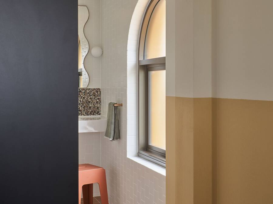 Een badkamer met een witte betegelde muur, een raam met een gebogen boog, een roze kruk en een handdoek aan een rek.