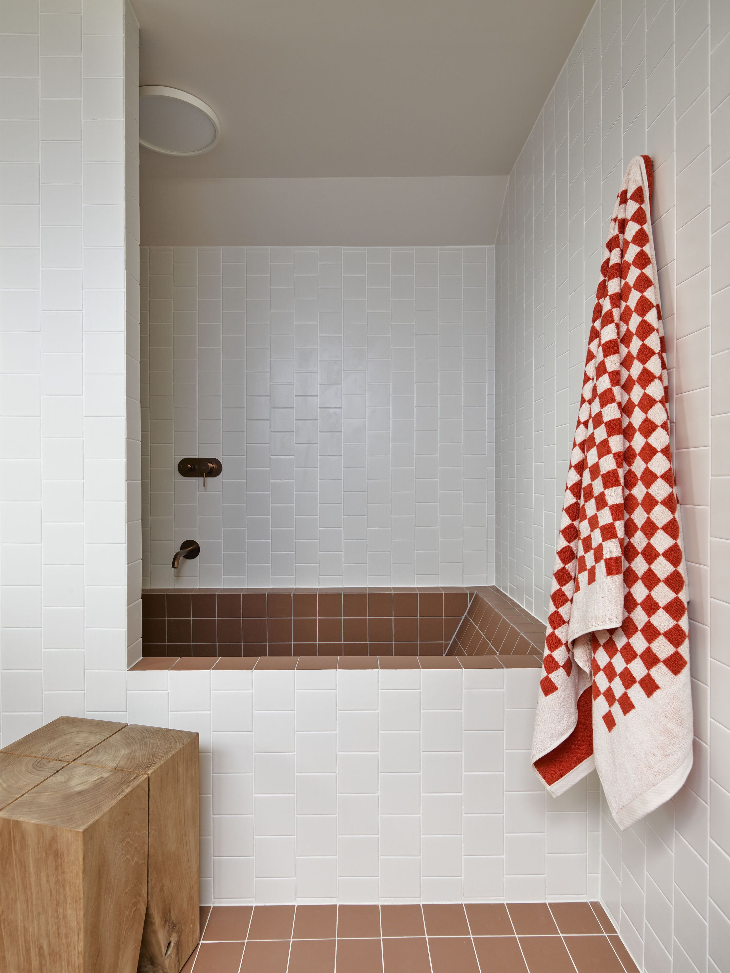 A bathroom with white tiled walls, a brown-tiled bathtub, a wooden bench, and a red and white checkered towel hanging on the wall.