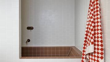 A bathroom with white tiled walls, a brown-tiled bathtub, a wooden bench, and a red and white checkered towel hanging on the wall.