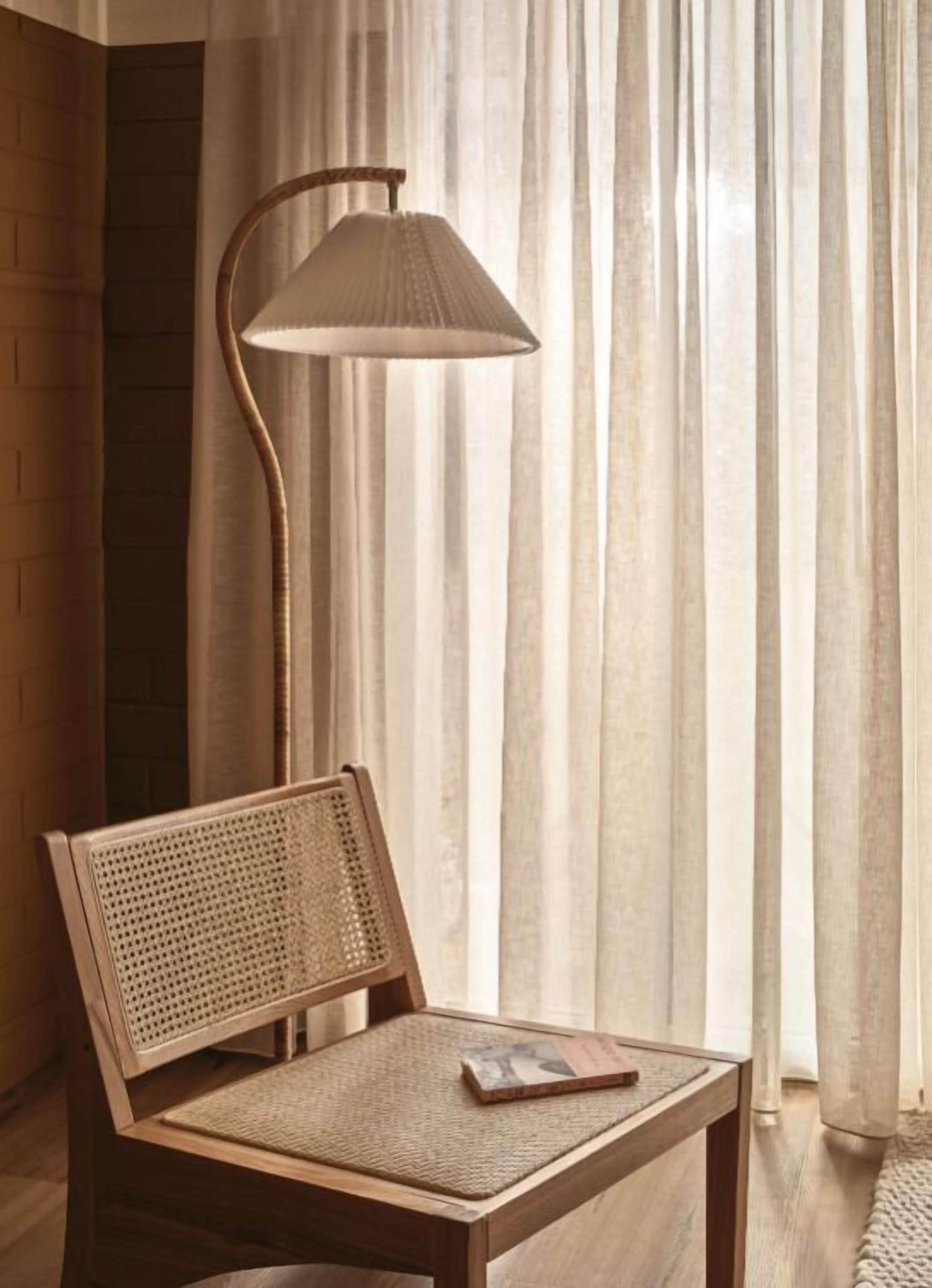 Una sedia di legno con schienale intrecciato e un tavolo, posti di fronte a una lampada da terra con paralume conico, su uno sfondo di tende leggere e un muro in mattoni.
