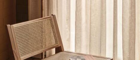 A wooden chair with a woven backrest and a table, positioned in front of a floor lamp with a conical shade, against a backdrop of sheer curtains and a brick wall.