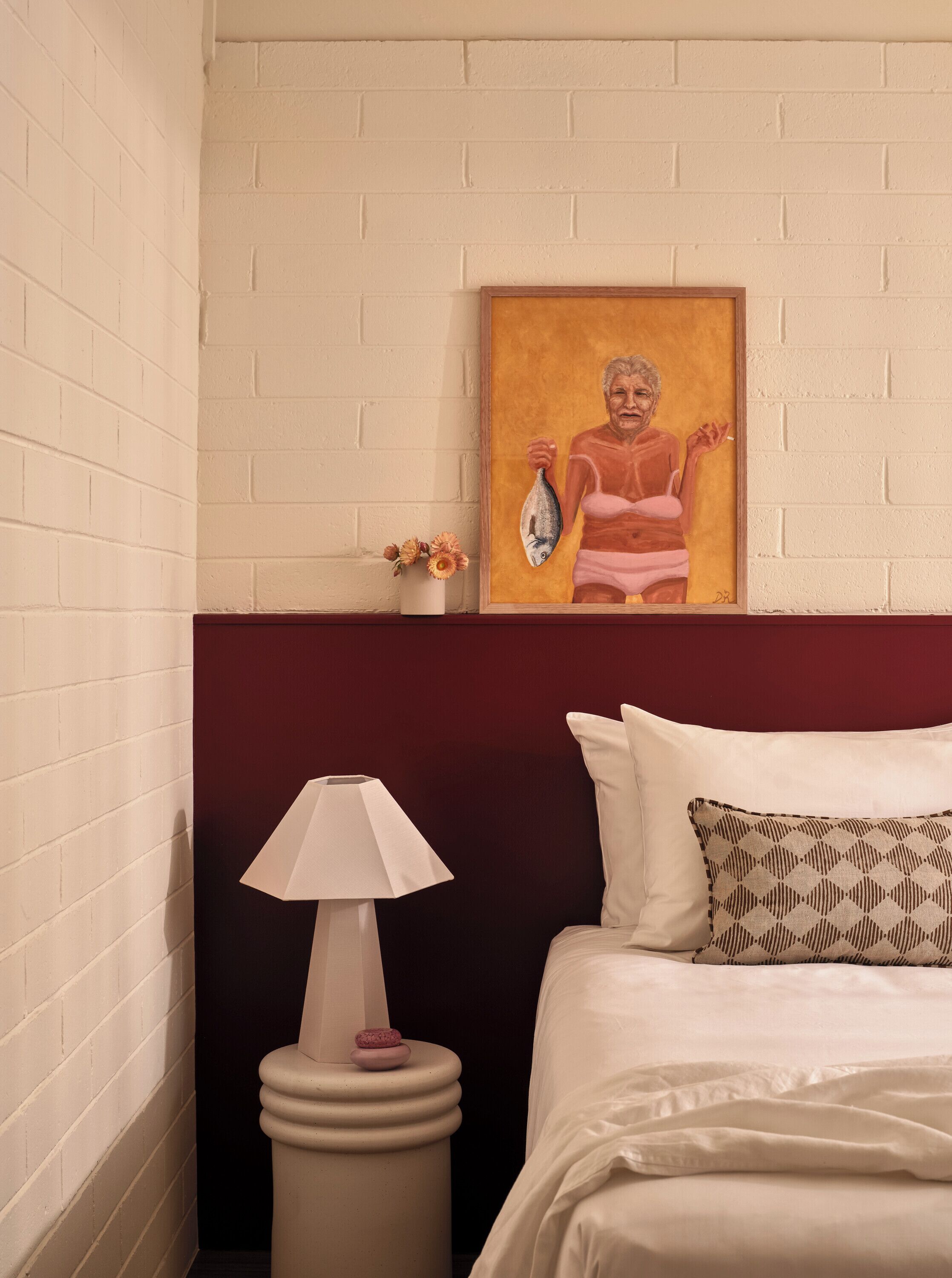 Een hotelkamer met een witte bakstenen muur, een schilderij van een persoon die een vis vasthoudt, een nachtkastje met een lamp en een bed met wit beddengoed en gestreepte kussens.