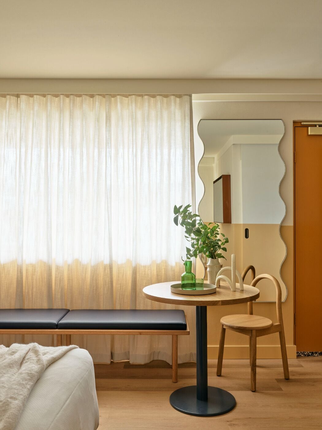 A hotel room with a bed, a round table with a green bottle and a plant, a bench, and a mirror with a wavy frame.
