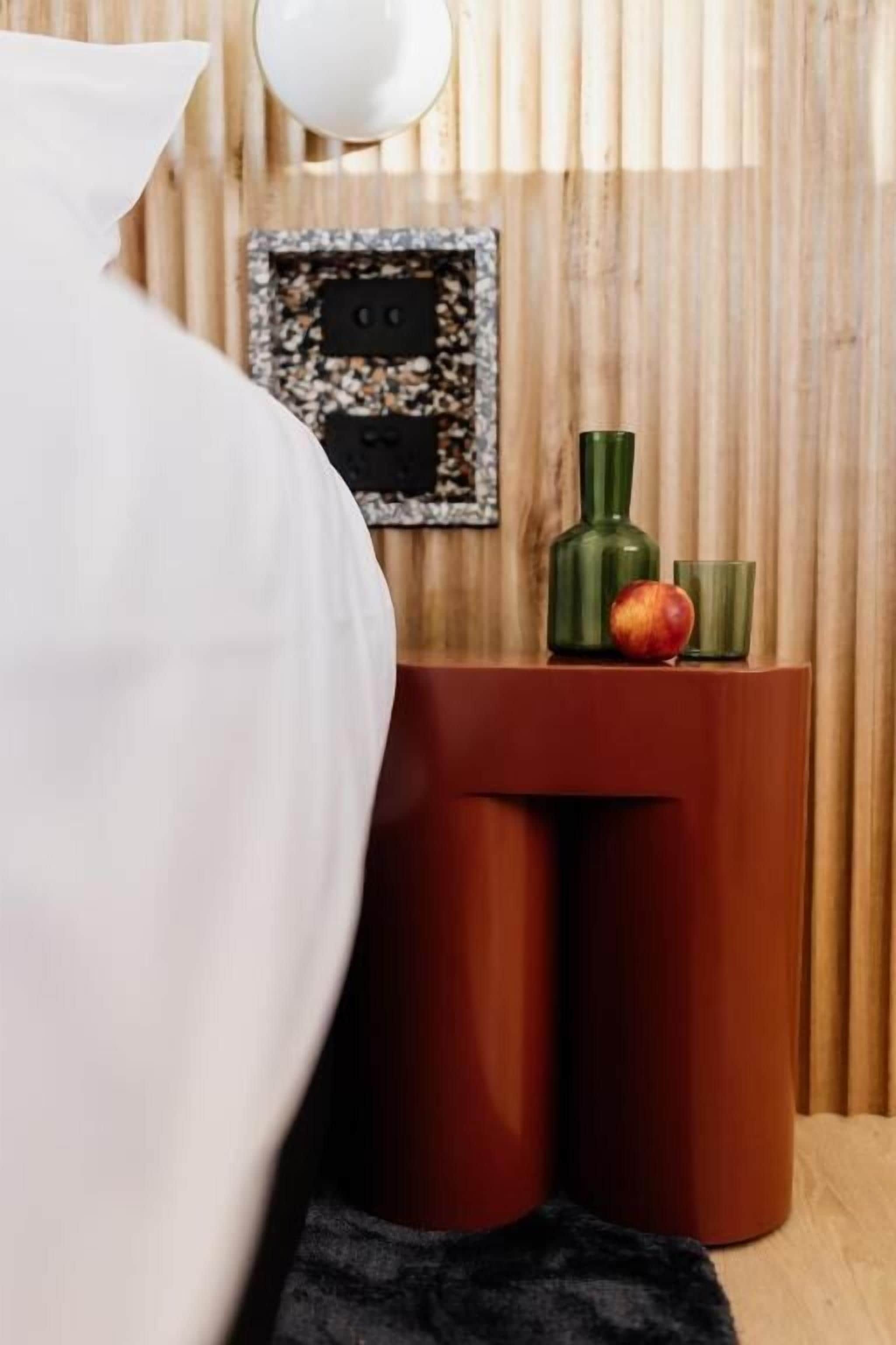 A bed with a white pillow and a dark throw blanket. In the background, there is a wooden wall with vertical slats, a framed picture, and a small table with a green vase and a fruit.