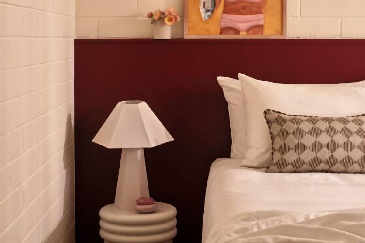 Habitación de hotel con pared de ladrillo blanco, un cuadro enmarcado sobre la cama, una lámpara de noche y un cojín estampado.