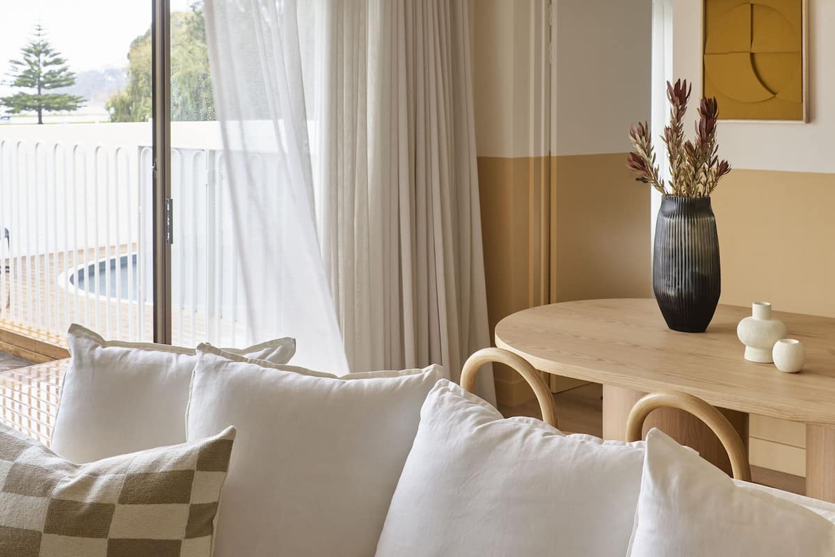 Habitación de hotel con sofá, una mesa de comedor, un jarrón con flores secas y vistas al exterior.