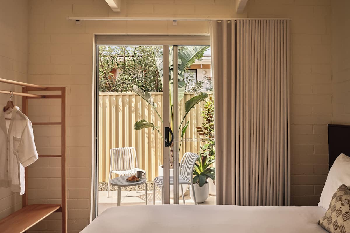Un dormitorio con cama, un perchero, una mesita con un plato y una taza, una silla y una planta en maceta.