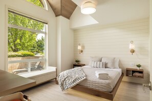 Un dormitorio con un ventanal grande, una cama con almohadas, mesitas de noche y un banco junto a la ventana.