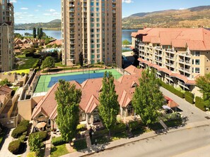 Un edificio de gran altura con cancha de tenis, rodeado de edificios residenciales y un lago.