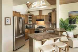 Una cocina moderna con refrigerador de acero inoxidable, gabinetes de madera, horno empotrado y una isla de granito con taburetes de bar.