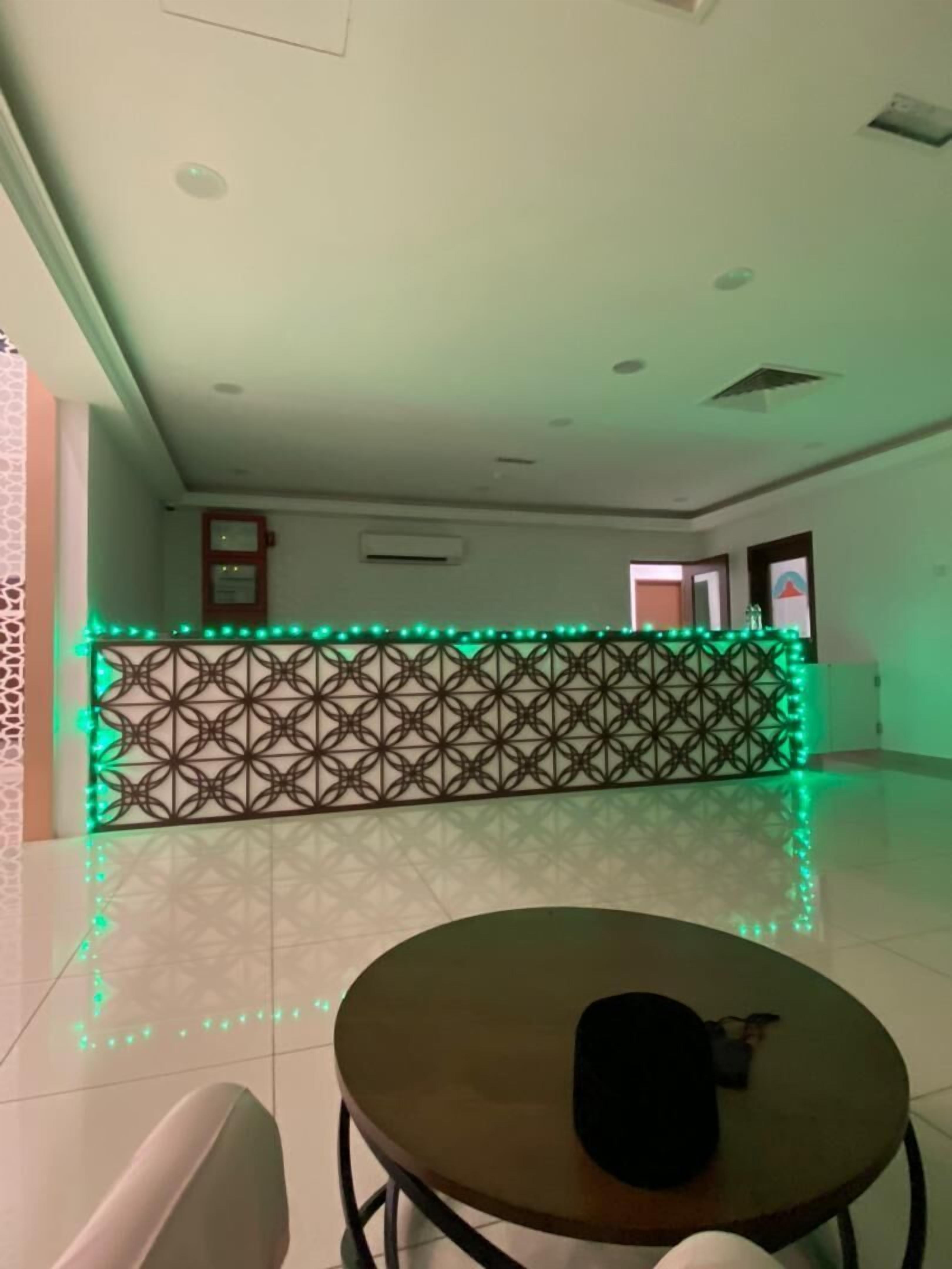 Un hall d’hôtel avec un paravent à motifs, une table ronde sur laquelle se trouve un objet noir, et un éclairage d’ambiance vert.