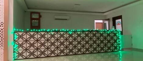 Um saguão de hotel com uma divisória estampada, uma mesa redonda com um objeto preto sobre ela e iluminação ambiente verde.