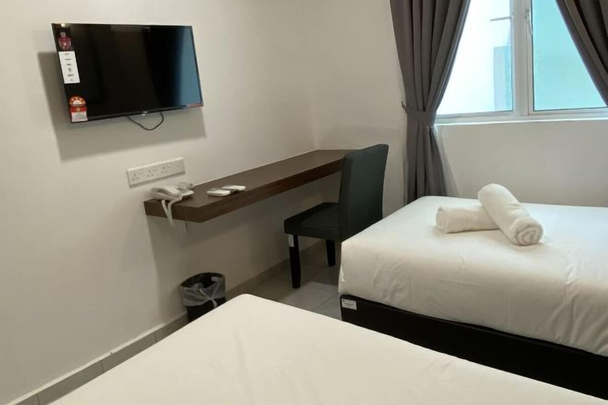 A hotel room with a bed, a desk with a chair, a TV mounted on the wall, and a window with curtains.
