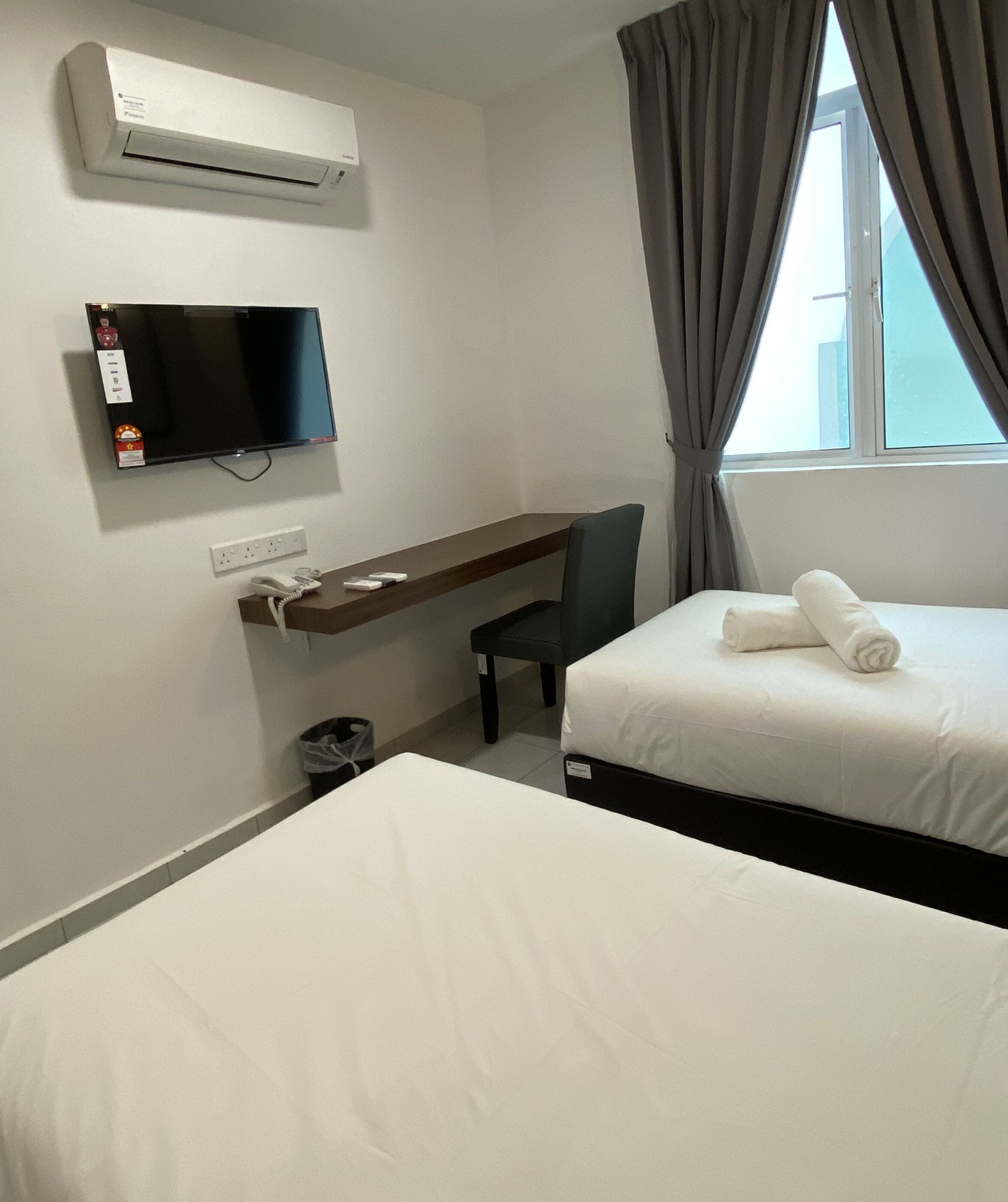 Quarto de hotel com duas camas, uma mesa com cadeira, televisão, aparelho de ar condicionado e janela com cortinas.