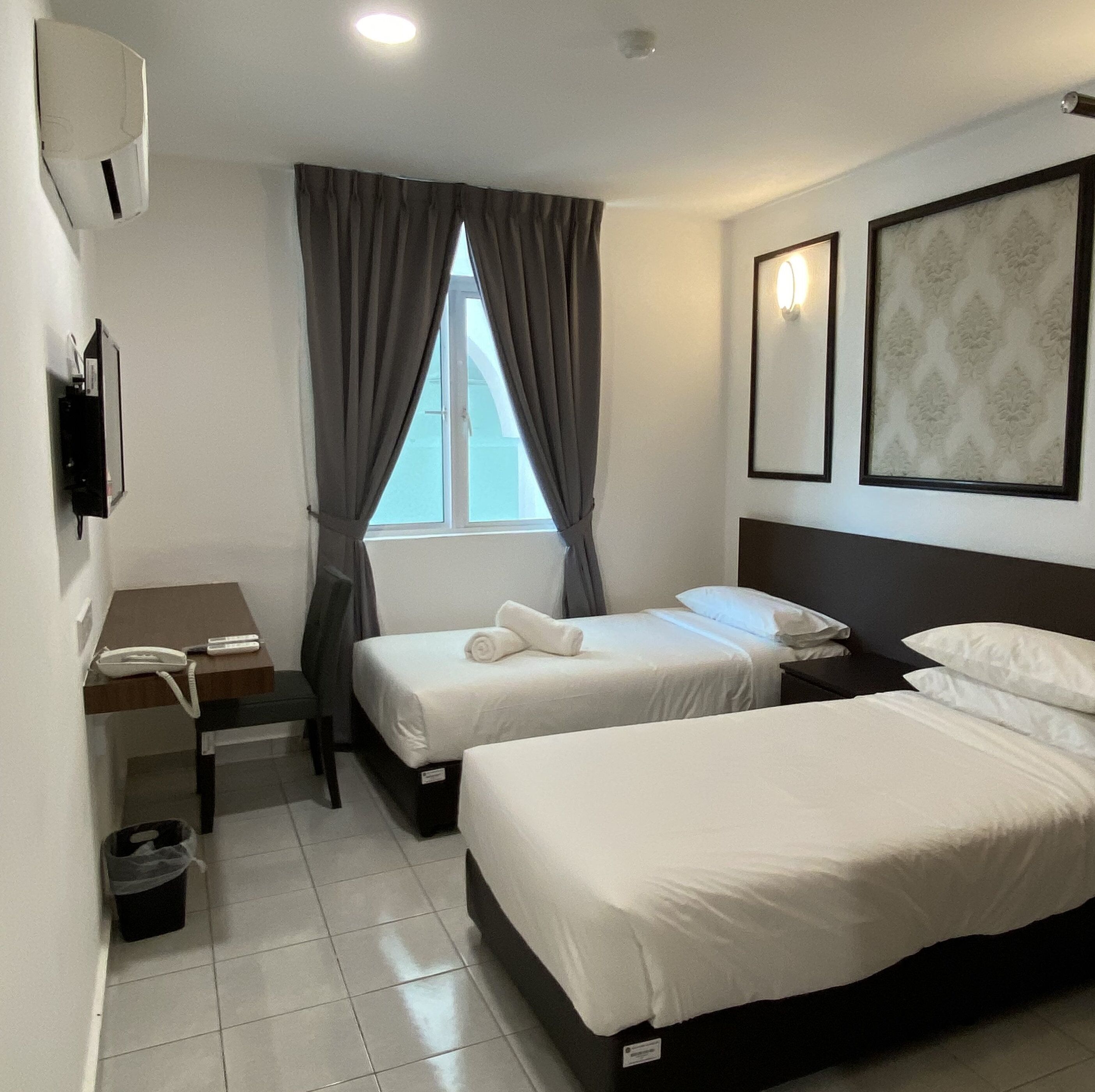 A hotel room with two beds, a desk with a phone, a trash bin, and a window with curtains.