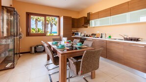 A modern kitchen with wooden cabinets, a dining table with chairs, and a view of palm trees through a window. - Harbour Views Gozitan Villa + Shared Pool, Mġarr Gozo, Malta (Mġarr Gozo)