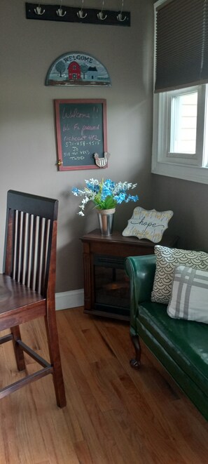Um quarto com uma cadeira de madeira, um sofá verde, uma pequena mesa com um vaso de flores, uma lousa com uma placa de boas-vindas e uma janela com persianas.